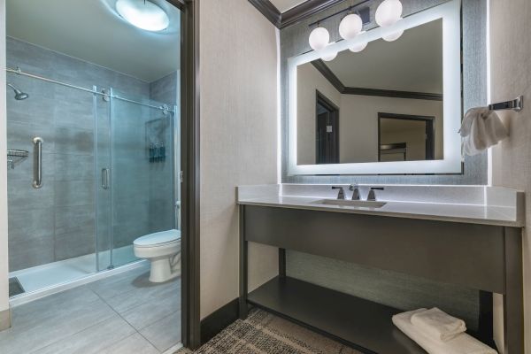 A modern bathroom with a glass shower, toilet, large mirror, a sink with lighting, and neatly folded towels on a shelf.