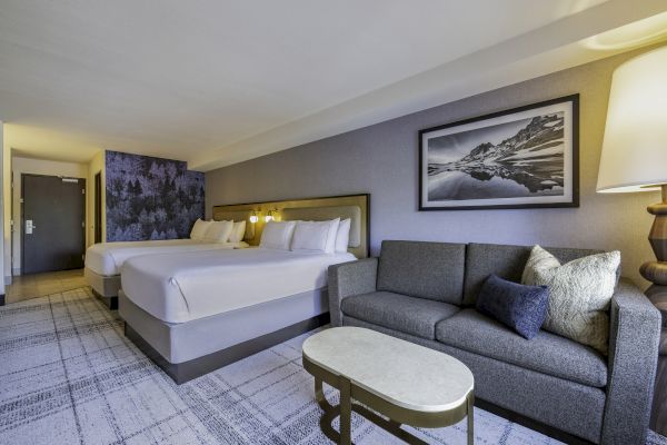 The image shows a hotel room with two beds, a gray sofa, a coffee table, and a framed mountain landscape picture on the wall.