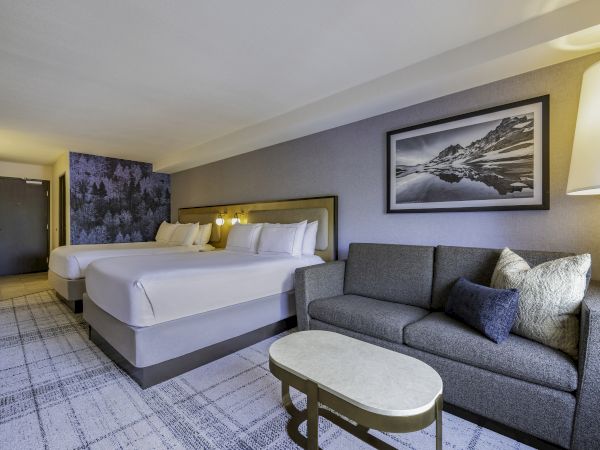 The image shows a hotel room with two beds, a gray sofa, a coffee table, and a framed mountain landscape picture on the wall.