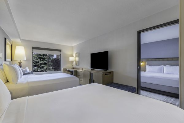 A modern hotel room with two beds, desk, TV, and window view, connected to another room with a bed through an open doorway.