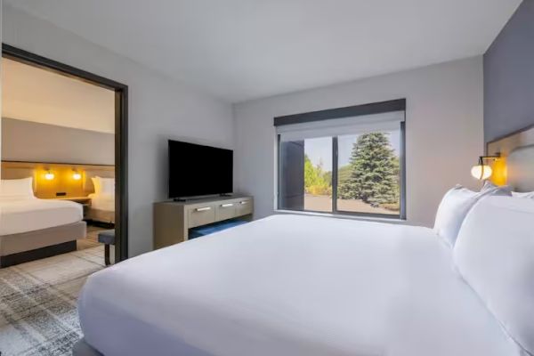 The image shows a hotel room with a bed, TV, window with a view, and an adjoining room visible through an open door.
