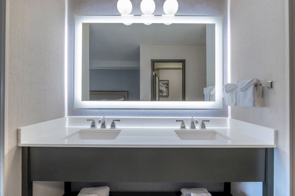 A bathroom vanity with a large lit mirror, dual sinks, and modern fixtures. Towel rack with neatly folded towels on the right.