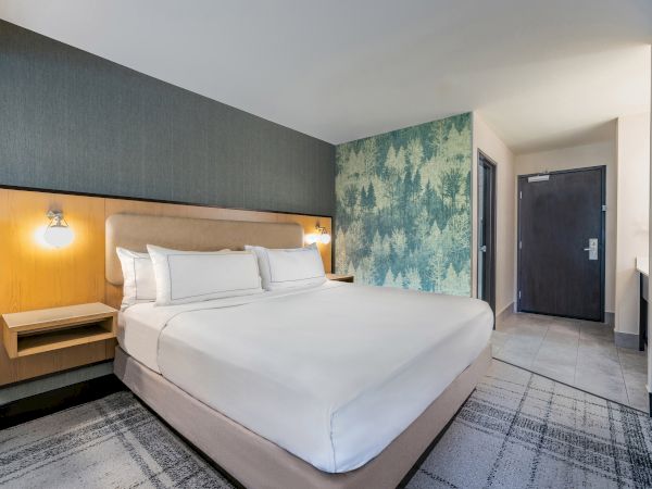 A hotel room featuring a neatly made bed, modern décor, wall lamps, forest-themed wallpaper, and a dark wooden door.