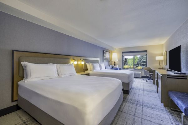 A modern hotel room with two large beds, a TV, desk, lamps, and a window showcasing greenery, creating a cozy and inviting atmosphere.