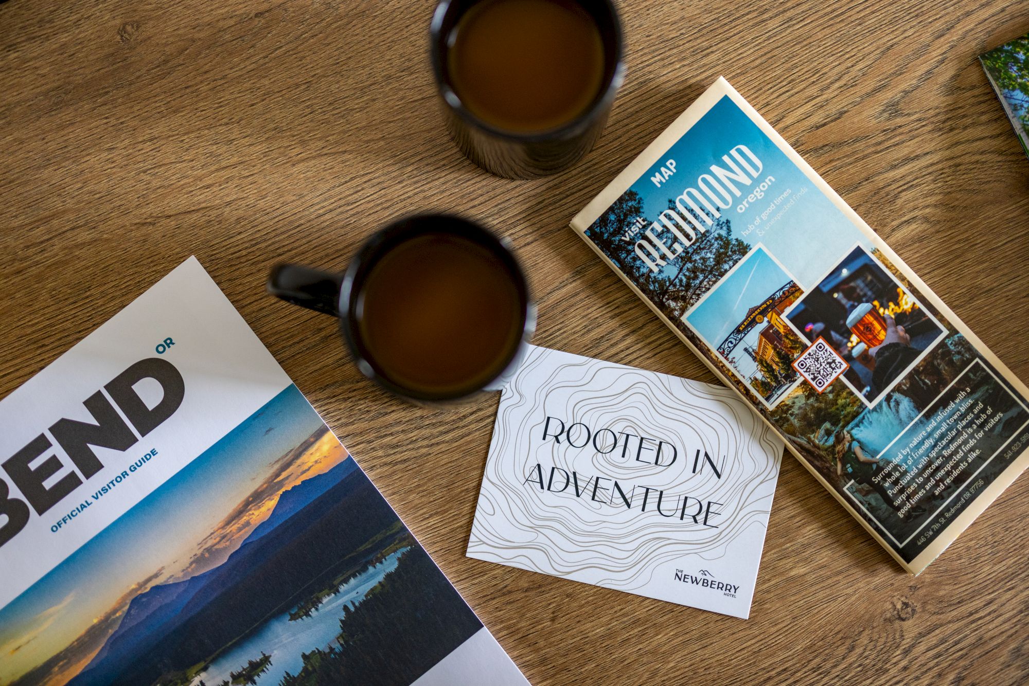 Coffee mugs, travel magazines, and a &ldquo;Born to Adventure&rdquo; card on a wooden table, with a scenic bend/landscape vibe.