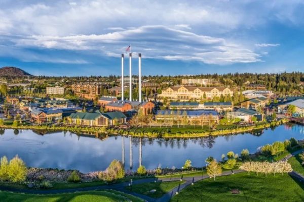 The image shows a scenic view of a lakeside complex with buildings, tall chimneys, and green spaces under a partly cloudy sky.