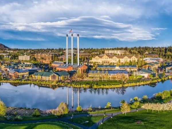 The image shows a scenic view of a lakeside complex with buildings, tall chimneys, and green spaces under a partly cloudy sky.