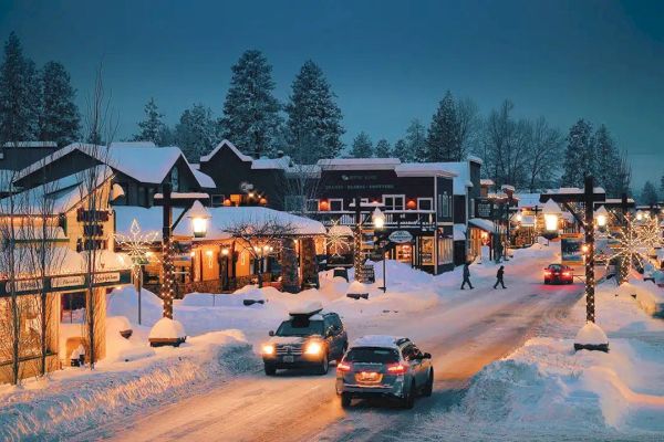 A snowy street scene with cars, decorated buildings, and trees, creating a cozy winter atmosphere under evening lights.