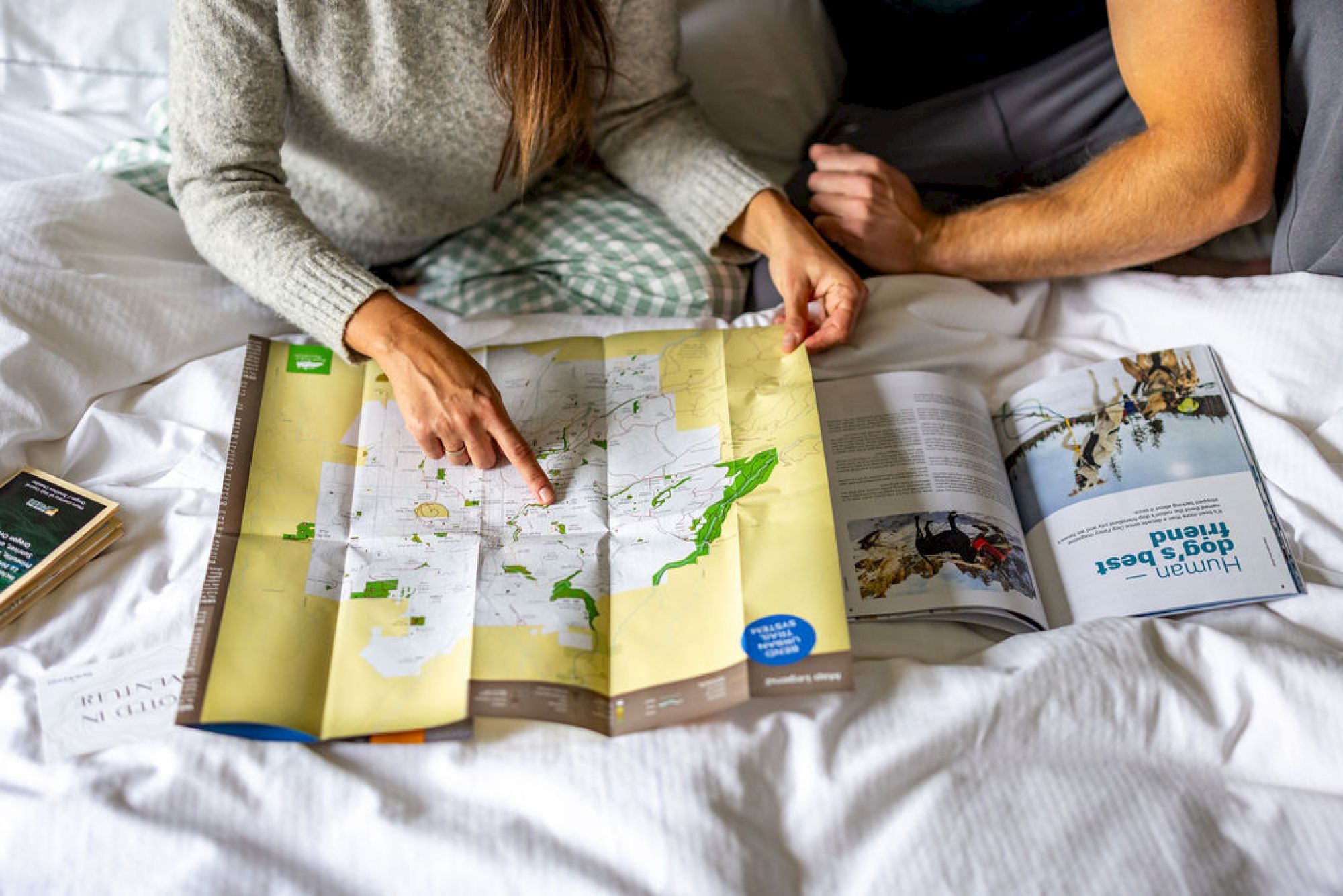 Two people sit on a bed, studying a large folded world map while a travel book lies open beside them, planning a trip together.