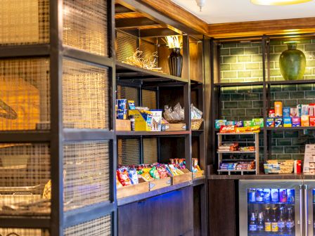 A small shop or pantry with dark wooden shelves, assorted snacks, beverages, and pantry items, plus a compact fridge in the corner.