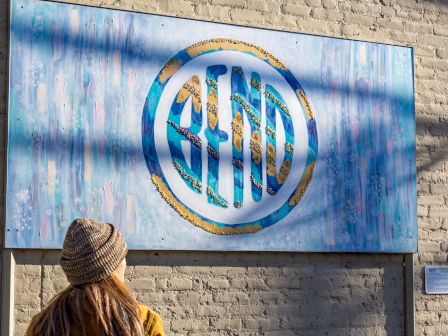 A person stands outside a weathered blue sign with a circular logo on a brick wall, wearing a yellow jacket and knit hat.