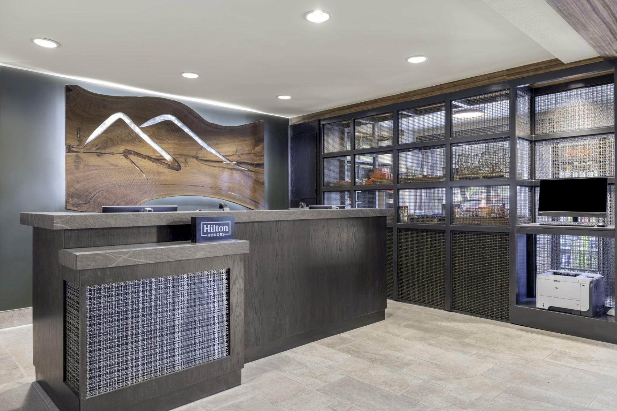 The image shows a hotel reception area with a modern design, featuring a wooden desk and a decorative wall. The shelf displays various items.