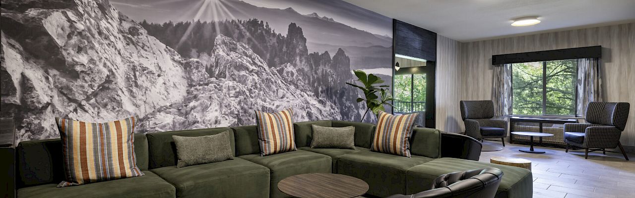A modern lounge area with a green sectional sofa, black chairs, round tables, and a large mountain mural on the wall.