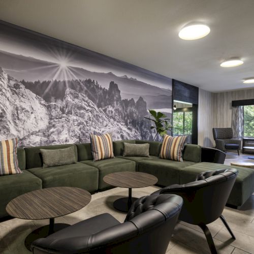 A modern lounge area with a green sectional sofa, black chairs, round tables, and a large mountain mural on the wall.