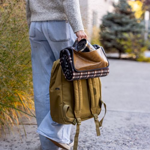 A person walks away carrying a tan backpack with a rolled blanket and a small dark bag tucked inside, on a sunny sidewalk.