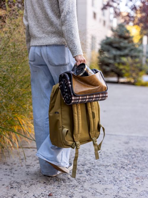 A person walks away carrying a tan backpack with a rolled blanket and a small dark bag tucked inside, on a sunny sidewalk.