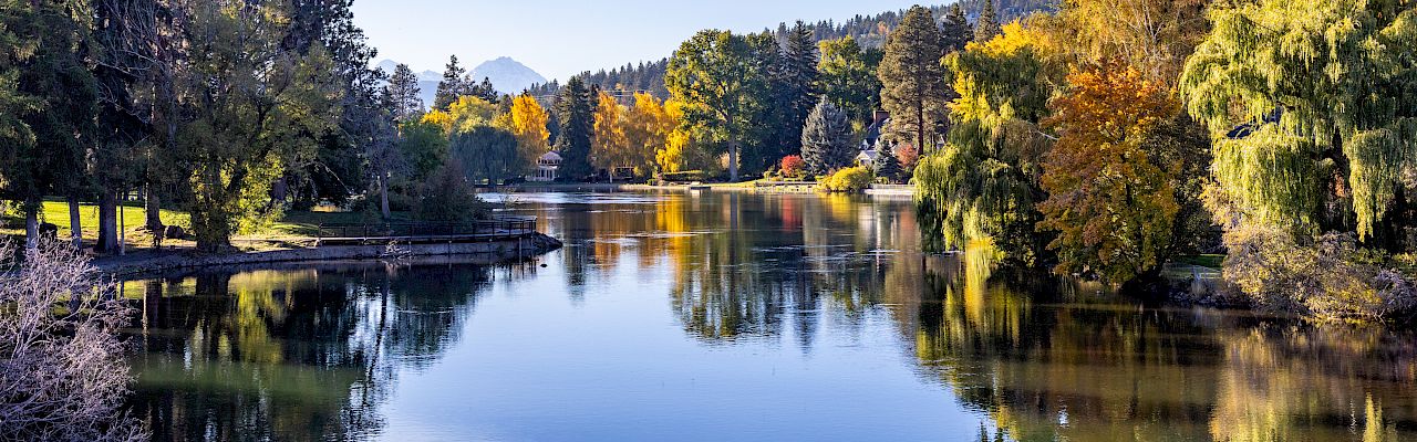 A tranquil lake mirrors the autumn trees in yellows and greens, framed by a clear blue sky and distant hills.
