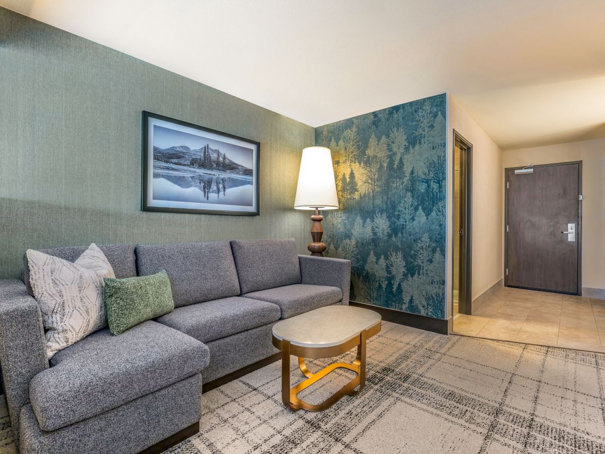 A modern living room with a gray sofa, decorative pillows, a floor lamp, artwork, and a round table on a patterned carpet.