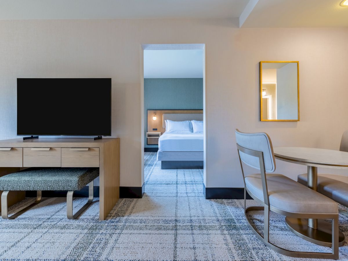 A hotel room features a TV, table, chairs, and a cozy bed visible through a doorway, with modern decor and a carpeted floor.