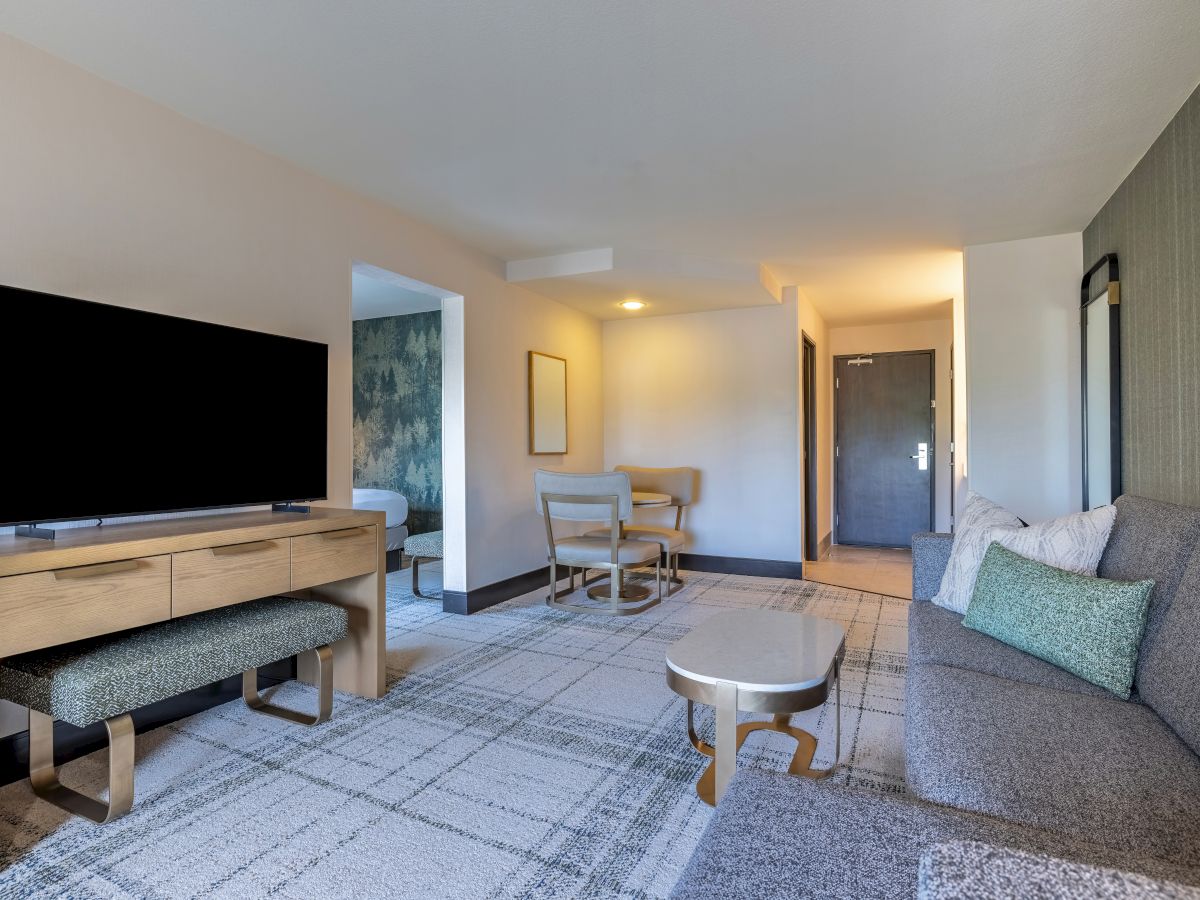 A modern living area with a sofa, TV on a wooden stand, small coffee table, and a dining area in the background, all with neutral tones.