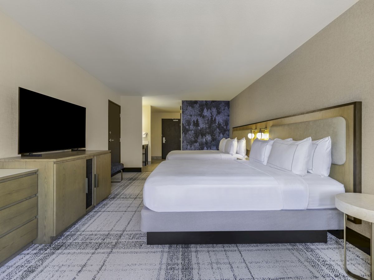 The image shows a modern hotel room with two beds, a flat-screen TV, and stylish decor on the wall.