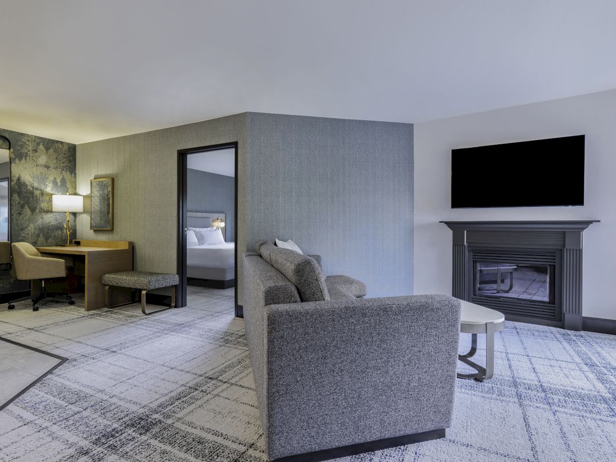 A modern living area with a sofa, TV above a fireplace, a desk with a chair, and a partial view of a bedroom.