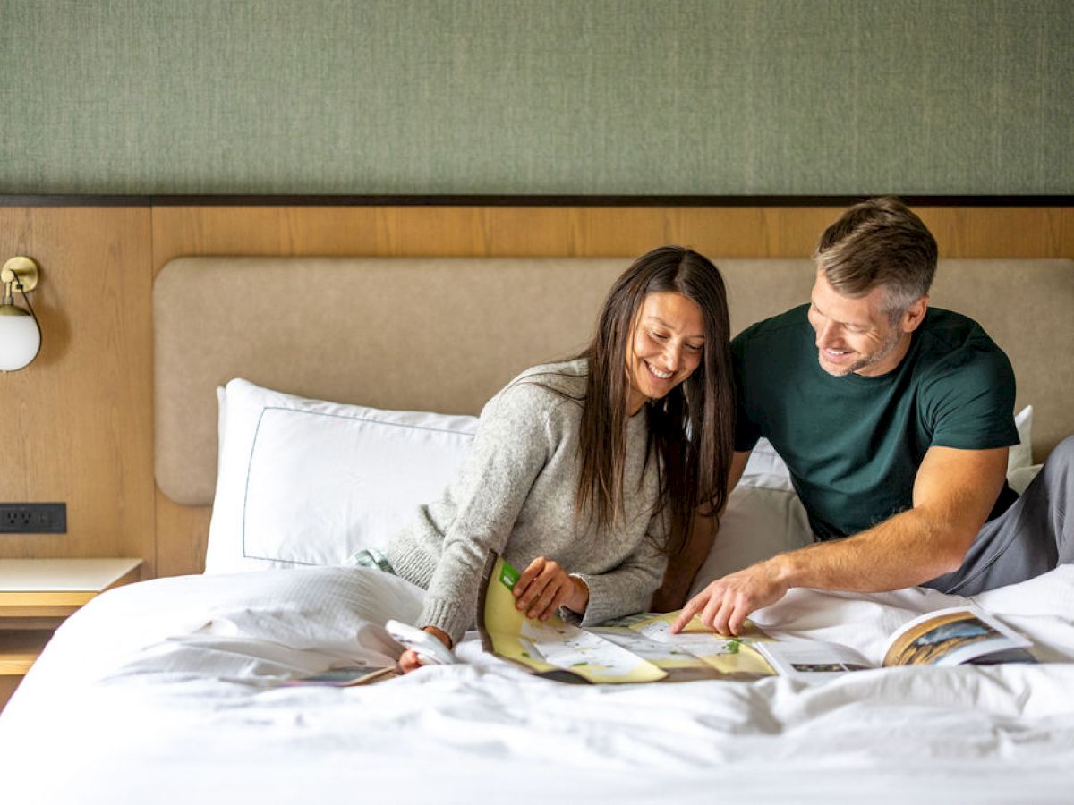 A couple sits up in bed looking at a book together, smiling and pointing at pages, enjoying a cozy morning moment.