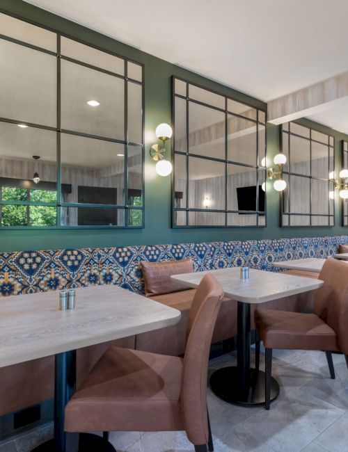 The image shows a modern dining area with tables, upholstered benches, decorative mirrors, and stylish lighting fixtures on the walls.