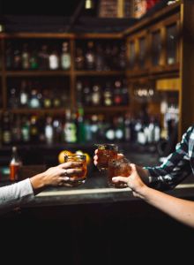 Three people are clinking glasses in a bar setting, with bottles blurred in the background.