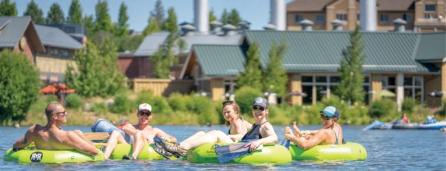 Five people are floating on inner tubes in a calm body of water near buildings and trees, enjoying a sunny day.