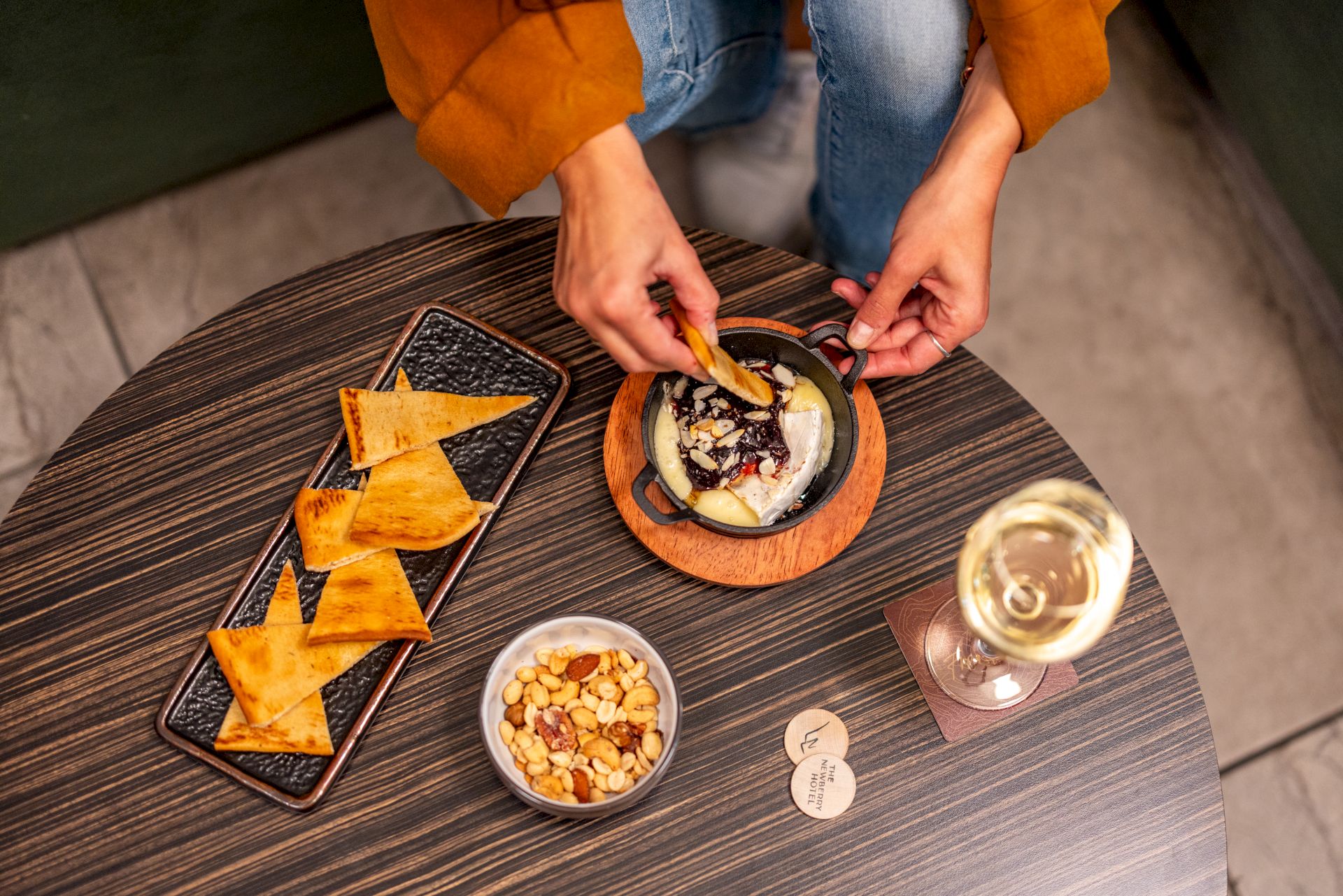 A person dips a tortilla chip into a dip on a round wooden table, with a tray of chips, a small bowl of nuts, a glass of white wine, and a few coin-like tokens.