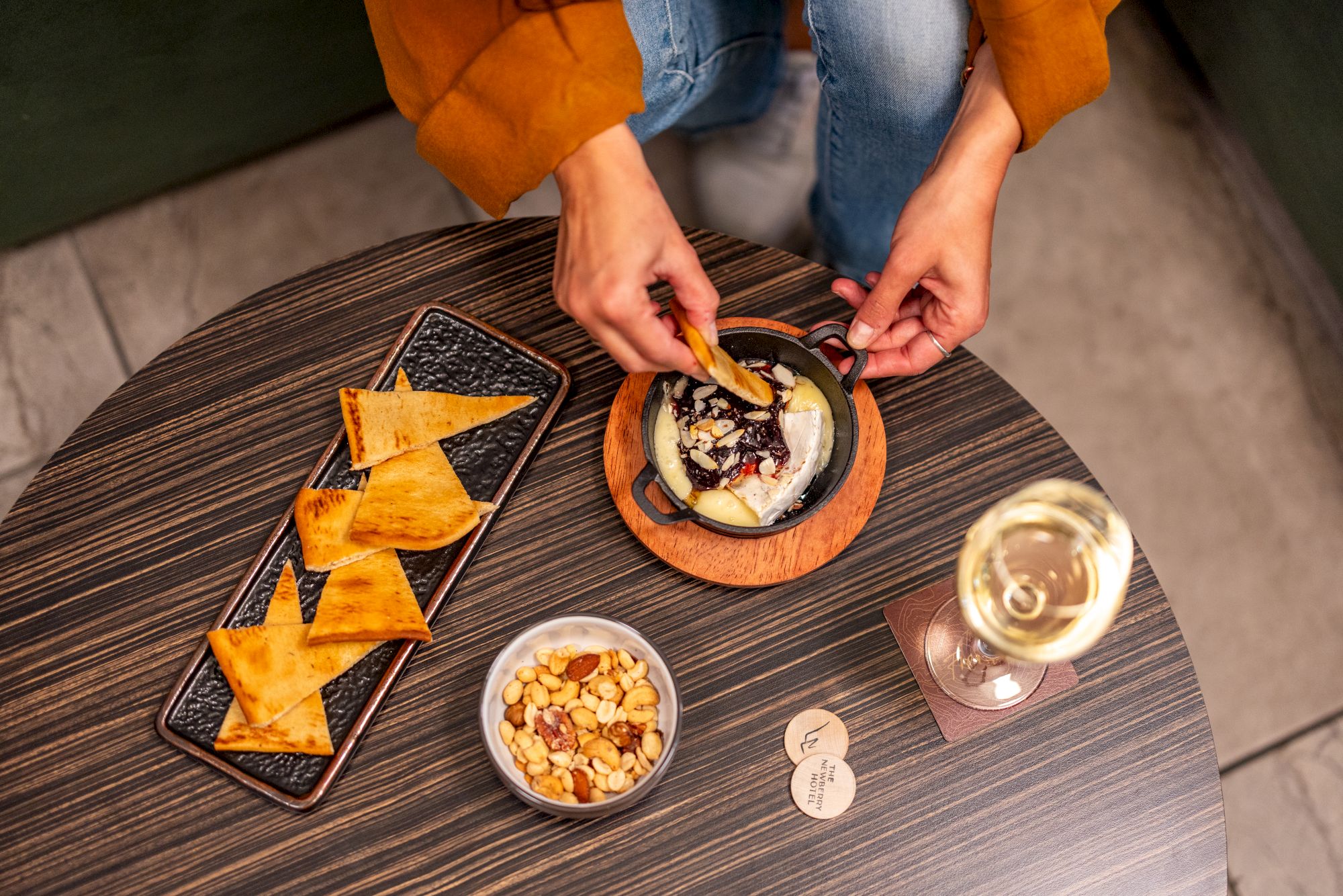 A person dips a tortilla chip into a dip on a round wooden table, with a tray of chips, a small bowl of nuts, a glass of white wine, and a few coin-like tokens.