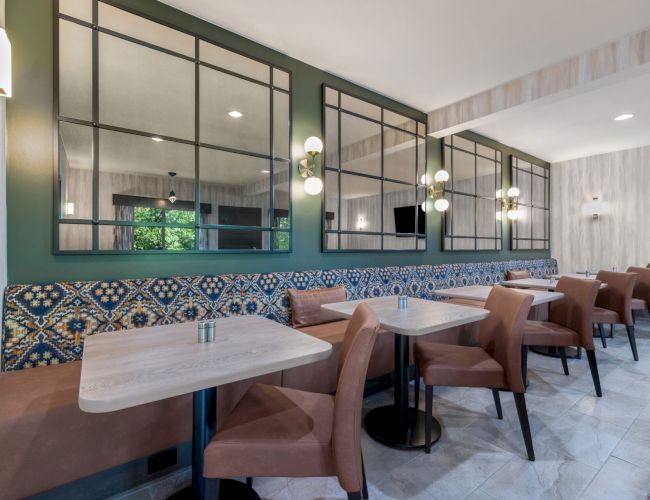 The image shows a modern dining area with mirrored walls, patterned seating, and arranged tables and chairs in soft earthy tones.