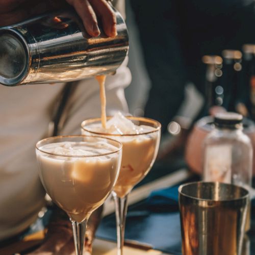 A person is pouring a creamy cocktail from a shaker into two glasses with ice, surrounded by bottles and bar tools.