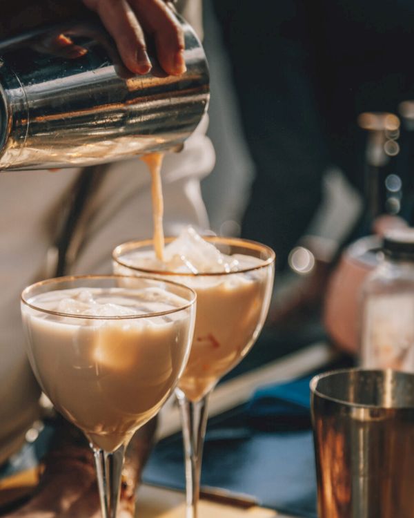 A person is pouring a creamy cocktail from a shaker into two glasses with ice, surrounded by bottles and bar tools.