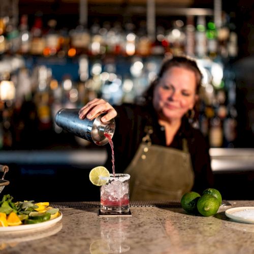 A bartender pours a drink with a lime wedge into a glass at a polished bar; fresh fruit and ingredients sit on the counter.