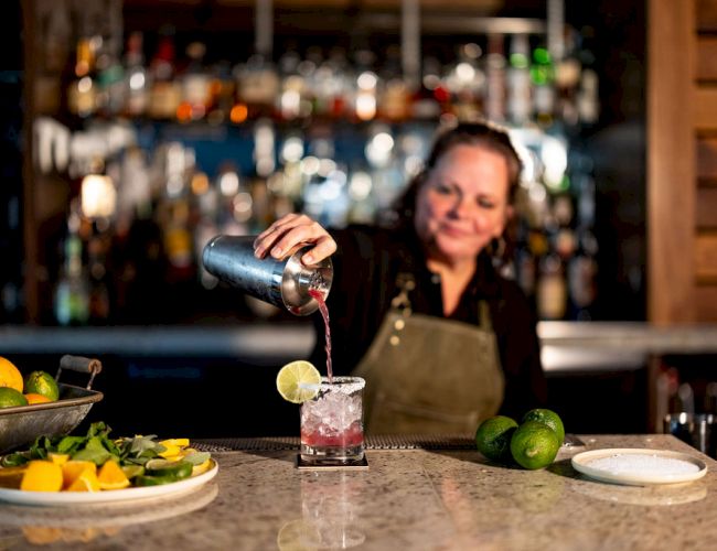 A bartender pours a drink with a lime wedge into a glass at a polished bar; fresh fruit and ingredients sit on the counter.