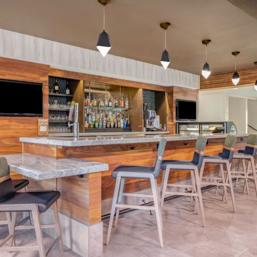 A modern bar with wooden accents, high chairs, pendant lights, a TV, and a variety of liquor bottles displayed behind the counter.