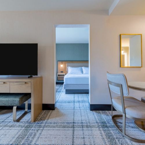 A hotel room with a TV, seating area, and a glimpse into a bedroom; neutral tones and modern furnishings create a cozy ambiance.