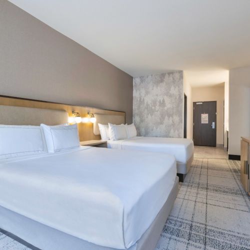 The image shows a modern hotel room with two neatly made double beds, a TV, and a doorway in the background.