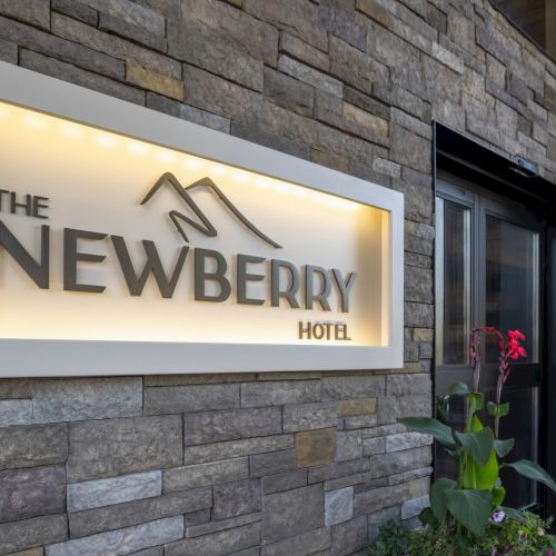 The image shows the exterior of The Newberry Hotel with a stone wall and illuminated sign, alongside some green plants in front.