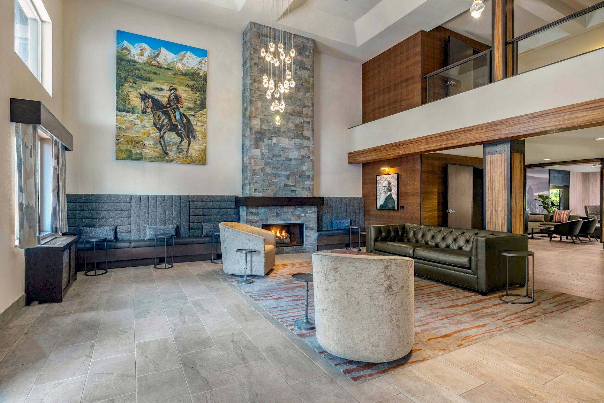 Spacious hotel lobby with mid-century seating, stone fireplace, modern chandelier, art on the wall, and a mezzanine loft above.