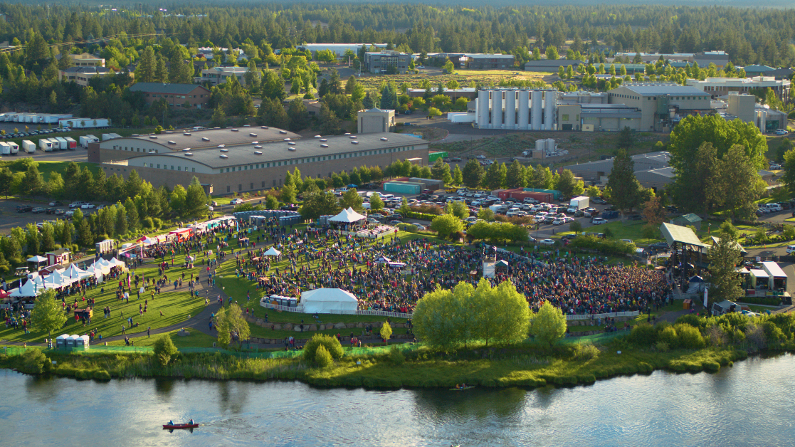 An outdoor concert event with a large crowd, tents, and a stage near a river, surrounded by buildings and trees in an industrial area.