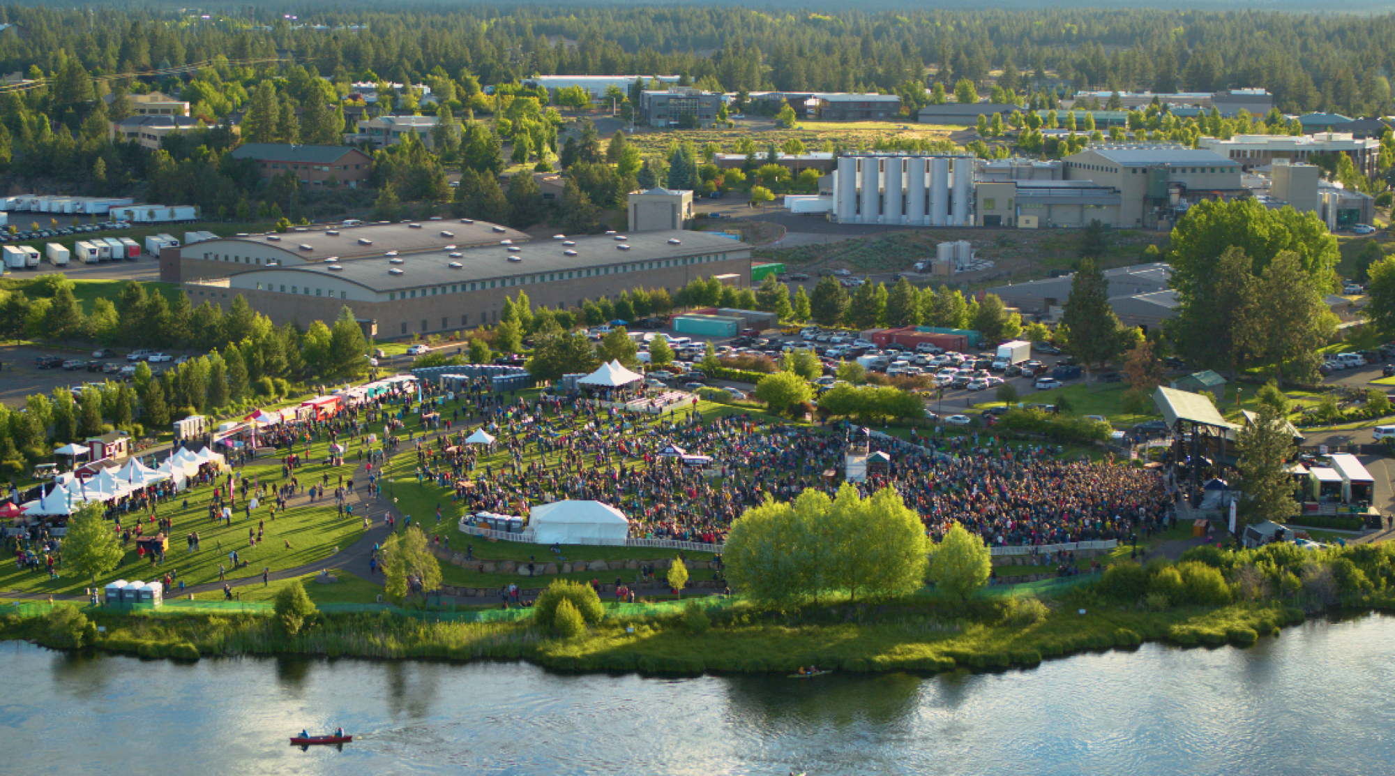 An outdoor concert event with a large crowd, tents, and a stage near a river, surrounded by buildings and trees in an industrial area.