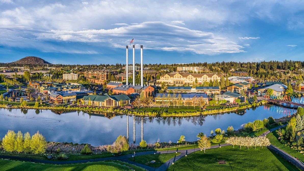 A scenic view featuring a lakeside park, buildings, three tall smokestacks, and lush greenery under a partly cloudy sky.