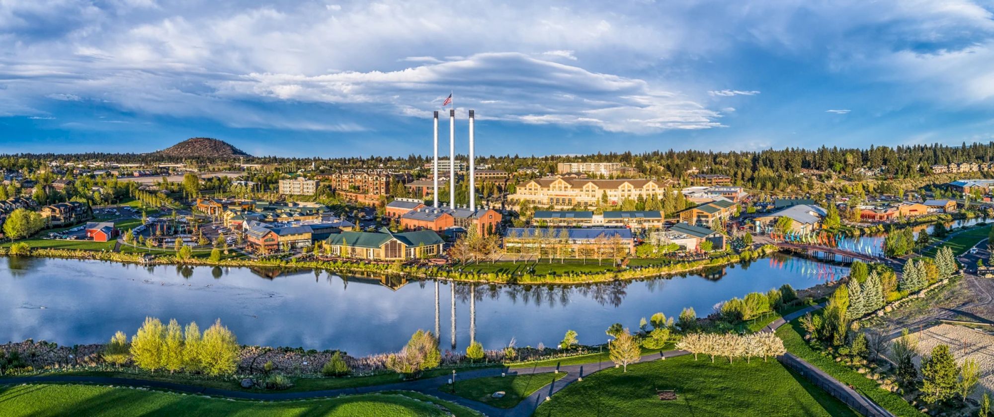 A scenic view featuring a lakeside park, buildings, three tall smokestacks, and lush greenery under a partly cloudy sky.