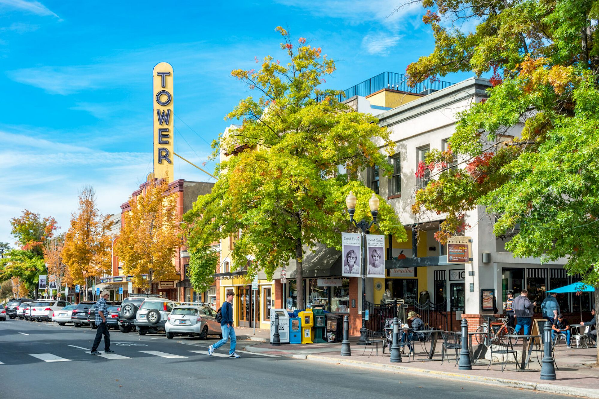 A vibrant street scene with shops, people, parked cars, and a sign reading "TOWER" amidst trees on a sunny day.