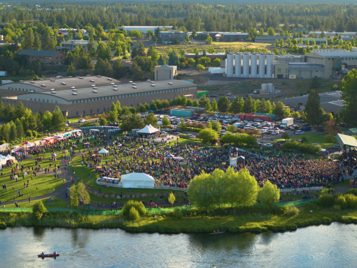 An outdoor concert with a large crowd surrounded by trees and industrial buildings near a river.