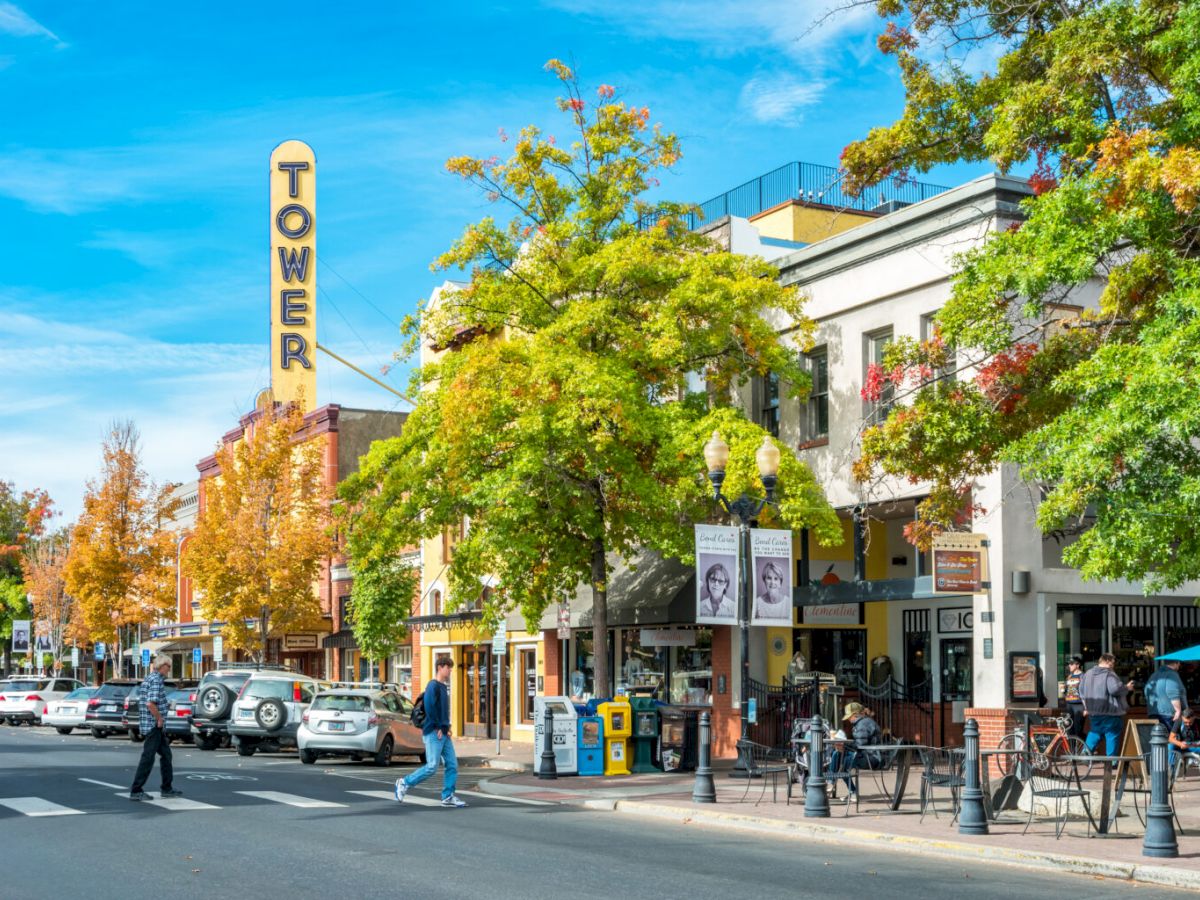 A vibrant street scene with a "TOWER" sign, cars parked, people crossing, and colorful trees lining the sidewalk with outdoor seating areas.