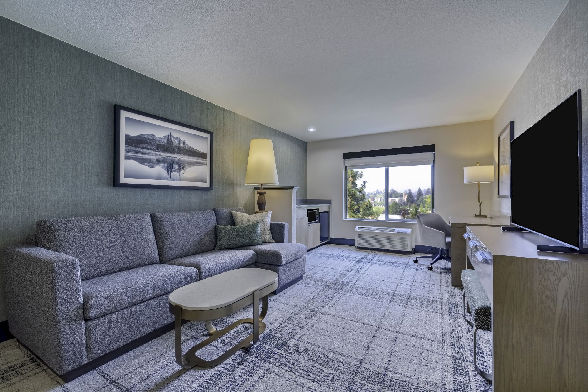 The image features a modern living room with a sofa, coffee table, TV, desk, and window with outdoor view.